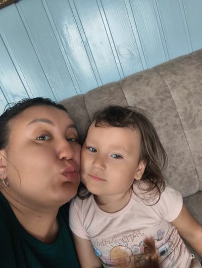 In this selfie, a woman with dark hair pulled back and wearing a dark green shirt is puckering her lips as if to kiss a young child. The child, with brown hair, is looking towards the camera with a slight smile, wearing a light pink t-shirt. They are both positioned on a textured, beige sofa. The background features a light blue paneled wall. The setting is consistent with a home interior. There is no indication of the city of Burayevo, Russia.