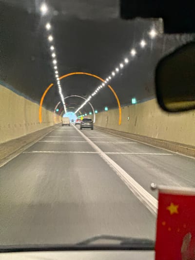 The image captures a forward-facing view from inside a moving vehicle within a multi-lane road tunnel in Xiehe, China. The tunnel features light-colored concrete walls and a dark ceiling illuminated by sequential white strip lights forming an arc overhead and extending down the tunnel. Orange-lit archways are visible at intervals, complementing the primary lighting. Green rectangular lights are positioned along the upper walls. The asphalt road surface displays clear white lane markings. Ahead, a white utility van, a dark passenger car, and a black SUV or sedan are visible, all traveling in the same direction. A portion of a rearview mirror is visible in the upper right, and a small red flag with a yellow star, consistent with the flag of the People's Republic of China, is partially visible in the bottom right corner, suggesting its attachment to the vehicle's interior.