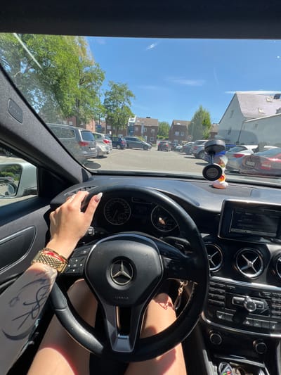 A first-person perspective shot shows the interior of a Mercedes-Benz, parked in an outdoor parking lot in Heinsberg, Germany, on a sunny day. The driver's hand rests on the steering wheel, adorned with a gold bracelet. The car's dashboard and central console are visible, along with the infotainment screen displaying the text "HUSTLE". A novelty rubber duck is perched on the dashboard, attached to a suction cup mount. Outside the car, other vehicles are parked in a line, and residential buildings and trees are visible in the background under a clear blue sky.