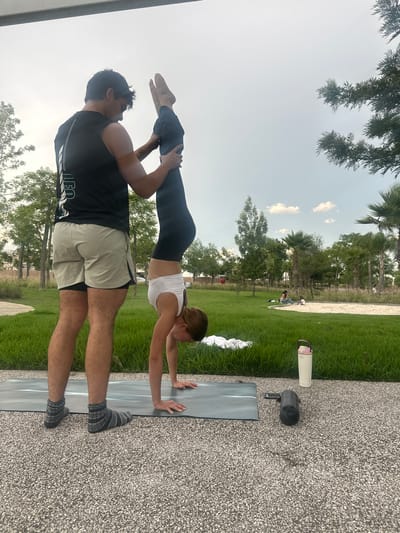 In the city of Tlaxcalancingo, Mexico, a man in a black tank top and tan shorts assists a woman performing a handstand on a yoga mat outdoors. The woman is upside down, with one leg extended vertically and the other bent at the knee. The man is positioned behind her, holding her extended leg for support. Both are on a paved area next to a grassy lawn. In the background, trees, palm trees, and a few other people are visible in a park-like setting. A white water bottle and a black cylindrical object are on the ground near the yoga mat.