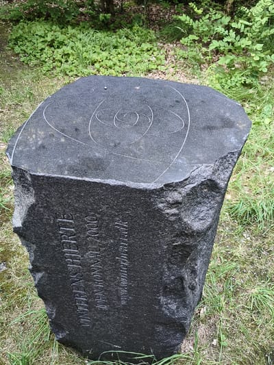 A rough-hewn, dark gray granite stone monument, approximately waist-high, is situated outdoors on a grassy area with some trees in the background. The top surface of the stone is relatively flat and inscribed with a series of concentric, oval-like lines, possibly representing orbits. The front-facing side of the stone is carved with text. The text appears to read "VINTERENS HJEMTE" vertically, followed by "JOHN RONNAU 2000" and a website address which is partially obscured. The monument is located in the city of True, Denmark.