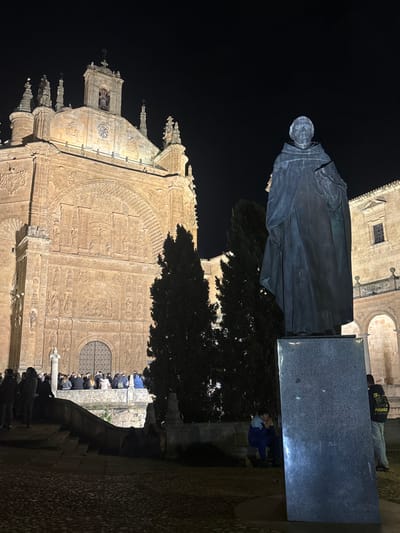 A nighttime scene captures the illuminated Plateresque facade of the Convento de San Esteban in Salamanca, Spain. In the mid-right foreground, a dark bronze statue of Fray Luis de Granada, depicted in a monastic habit and holding a book, stands on a dark, rectangular pedestal. The ground in the immediate foreground is paved with cobblestones.

In the background, the elaborately carved facade of the Convento de San Esteban church is visible, featuring an ornate main entrance, a large archway with relief sculptures, and a bell tower topped with a cross and pinnacles, all brightly lit against a dark, clear sky.

A group of approximately 15-20 individuals is gathered on a lower stone platform or wide steps directly in front of the convent's main entrance, partially obscured by a stone balustrade. They are mostly standing, appearing to observe the building. Two tall, dark evergreen trees are positioned between the statue and the convent's facade. To the far right, part of an adjacent building with arched cloisters is visible, also illuminated. Two additional individuals are present in the right foreground: one seated on the ground near the statue's base, and another standing further right, partially obscured, wearing a dark jacket with yellow text.
