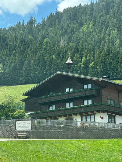 The image shows a wooden building with multiple stories and balconies, set against a backdrop of a dense forest and a green hillside. A sign in front of the building reads "Unterbaumgarten Urlaub am Bauernhof Familie Oberauer, Hochweg 9". The building has a traditional Alpine architectural style, with dark wood exterior and green trim on the balconies and roof edges. The balconies are adorned with white flowers. A small decorative turret with a weather vane sits on the roof. The foreground features a patch of green grass, a wooden bench, and a stone wall with a picket fence. In the distance, the forest covers the hillside, suggesting a rural or mountainous setting. The sky is partly cloudy. The location is identified as Reitdorf, Austria, Hochweg 9. There are no people or animals visible in the image.
