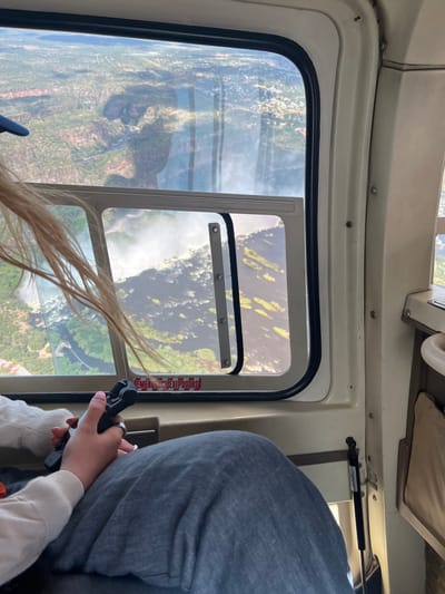 Inside a helicopter, a person is holding a handheld microphone in their right hand, a wedding ring is on their left ring finger. The window reveals the landscape of Victoria Falls Municipality, Zimbabwe. The landscape features a waterfall and river surrounded by green vegetation. The helicopter's interior includes a cream-colored frame around the window with a red emergency exit label.