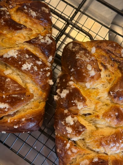 Two golden-brown, braided loaves, featuring a glossy crust and generously sprinkled with coarse white sugar crystals, are positioned side-by-side on a black wire cooling rack. The rack rests on a light-toned surface, which partially reflects light, within an unidentifiable indoor environment. No individuals are present. This scene is located in Bretton, United Kingdom.