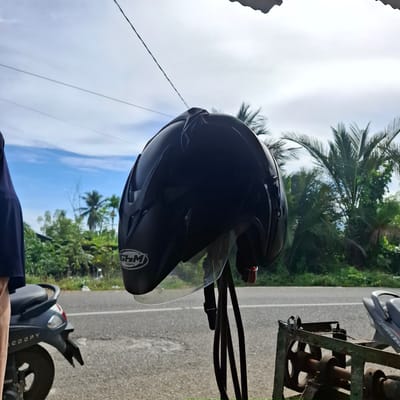 A black motorcycle helmet with a clear visor is hanging on a stand in front of a roadside scene. The helmet features a white logo with the letters "GHM" and the word "HELMETS" below it. To the left of the helmet, a person wearing a dark blue top and light-colored pants is partially visible, standing next to a dark grey scooter with "SCOOPY" written on its rear fender. The scooter is parked on a paved road with a white line marking. In the background, lush green vegetation and palm trees are visible under a bright, partly cloudy sky. The scene appears to be located in a tropical environment, likely in the vicinity of Rhieng Krueng, Indonesia, given the vegetation and scooter model. A rusty metal structure with a wheel is situated to the right of the helmet stand.