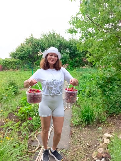 In Vyazovka, Russia, a woman in a white hat and white athletic wear stands in a garden, holding two clear plastic buckets filled with strawberries. The garden appears to be a backyard with abundant greenery and some visible produce plants. The woman is smiling and looking at the camera, holding one bucket in each hand at waist level. The background shows trees and a partially visible building, suggesting a rural or semi-rural setting. The ground is a mix of dirt, grass, and some stones, with a hose visible in the lower left corner. The sky is overcast.