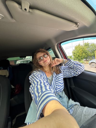 A young woman with blonde hair is taking a selfie in a car, with her right hand resting on her chin. She is wearing sunglasses, a blue and white striped shirt, and denim shorts. The interior of the car is visible, including the sun visor, seats, and parts of the dashboard. Outside the car, trees and a street are visible, suggesting a suburban or rural setting. The image appears to be taken during the daytime, with natural light illuminating the scene. The specific location of Kachimir, Russia, cannot be determined from the visual information provided in the image.