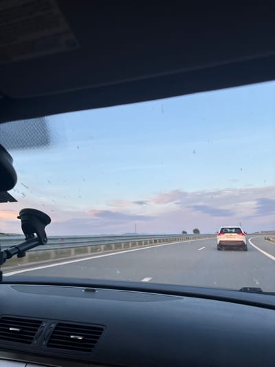 The image shows a view from inside a car driving on a highway in Aldomirovtsi, Bulgaria. A white car is visible in the distance ahead, traveling in the same direction. The sky is visible through the windshield, displaying a mix of blue and sunset colors. A suction cup mount is affixed to the windshield, holding a device that is partially visible. The car's dashboard and air vents are visible in the foreground. Roadside barriers are seen along the highway.