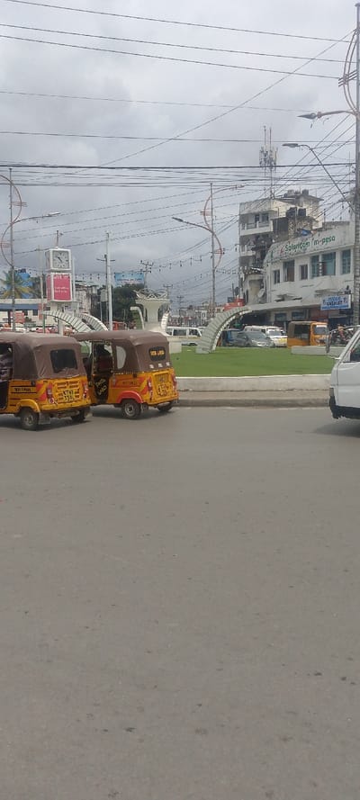 The image displays an urban street scene at an intersection within Majengo ward, Kenya, under an overcast sky. In the immediate foreground, a gray asphalt road occupies the lower portion of the frame. Two yellow and brown three-wheeled auto-rickshaws, commonly known as tuk-tuks, are visible on the left side of the road. The tuk-tuk closer to the camera on the left displays "TAXI 3 PASS" and license plate "KTMB 339L" on its rear; an occupant is partially visible within. The adjacent tuk-tuk exhibits "FATA LAXO" on its rear, also containing visible occupants.

A raised concrete curb separates the road from a central, grass-covered island. This island hosts a white, modern-style sculpture featuring multiple arches and a central decorative element. Several streetlights with distinctive orange spiral-shaped support structures are positioned around the island and along the thoroughfare. In the background, behind the tuk-tuks and the central island, multiple multi-story buildings are present. A prominent white building on the right side of the intersection features "Safaricom m-pesa" branding on its upper facade and a "FUSAM CHEMIST" sign below. Further back, a square clock tower with multiple faces is visible above other buildings. A tall communication tower is discernible above the distant structures. Numerous overhead electrical and communication lines crisscross the sky throughout the scene. Other vehicles, including cars and vans, are visible on the roads surrounding the central island. The partial rear of a white van is visible on the far right edge of the foreground road.