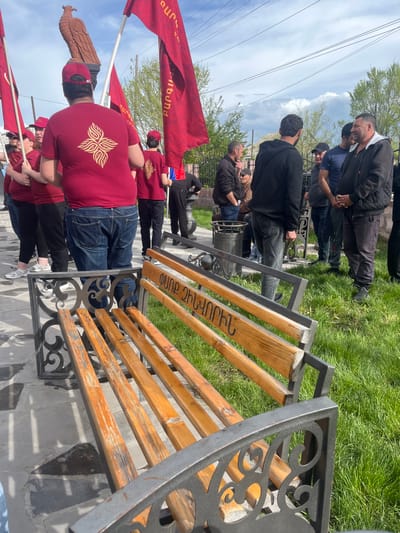 The image captures an outdoor scene, likely in the city of Akhuryan, Armenia, focusing on a group of people gathered near a bench and a statue. The bench is wooden with a metal frame, featuring decorative flourishes, and has Armenian inscriptions on its wooden slats. To the left of the bench, several people stand, many wearing matching burgundy T-shirts adorned with a gold-colored symbol on the back. Some hold red flags with what appear to be Armenian inscriptions as well. Further in the background, another group of people is standing on a grassy area near a trash can. The environmental backdrop includes trees, a cloudy sky, and power lines overhead. The statue to the left appears to be that of a bird with detailed feathers.