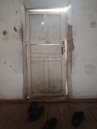 The image depicts an interior scene of what appears to be a room in Yusupov, Uzbekistan. A weathered, white wooden door stands prominently in the center, with light emanating from a gap at the top and along the side. The door is divided into multiple panels and has a dark metal handle. The walls are painted white but show signs of age, with peeling plaster and water stains.

In the foreground, a wooden floor is visible, laid with planks. Several pairs of black slip-on sandals are scattered on the floor. One pair is in the lower left, another in the lower right, and a third is slightly obscured in the center bottom. The lighting is dim, with the primary light source coming from the crack around the door, casting shadows on the floor and walls. The overall impression is one of a simple, somewhat neglected living space.