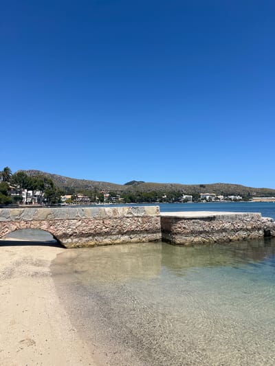 The image depicts a clear, sunny day at the Port De Pollença, Spain. In the foreground, a sandy beach meets the calm, clear water. A stone structure, likely a walkway or a small bridge, is partially submerged in the water. The stone construction features rough-hewn rocks of varying colors, including pinkish-red and grey, with some cement binding them together. The water near the shore is shallow and transparent, revealing the sandy and pebbly seabed. Further out, the water deepens to a vibrant blue.

In the mid-ground, a body of water stretches towards a coastline dotted with buildings and lush green trees. The architecture of the buildings appears to be Mediterranean in style, with white walls and red-tiled roofs. Behind the coastal town, a range of hills covered in sparse vegetation rises under a clear, brilliant blue sky. The overall scene conveys a serene and picturesque coastal landscape. There are no people visible in the frame.