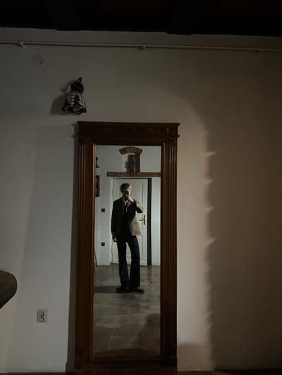 An indoor environment in Nałęczów, Poland, is depicted, centered on a tall, ornate wooden-framed mirror. Reflected within the mirror is an individual, appearing male, dressed in a brown tweed-patterned blazer, dark vest, collared shirt, tie, and dark flared trousers. The individual holds a black smartphone at chest height, obscuring their face, indicating the act of photographing their reflection. The background visible in the mirror includes a white door, a wall with an arched niche containing a small sculpture or decorative object, and dark, patterned floor tiles. To the left of the mirror, a white wall features a decorative wall lamp with a bell-shaped, multi-colored glass shade and a white double electrical outlet near the floor. The upper wall displays a faint circular mark. Above, a dark ceiling beam and a light-colored pipe are present. The white wall to the right of the mirror exhibits multiple distinct, vertical, jagged shadows. A dark, curved object is partially visible on the far left edge of the frame. The scene is dimly lit.