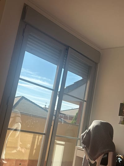 The image captures an interior view looking outwards through a multi-pane sliding glass door with grey frames, located within a residential property in Paracuellos de Jarama, Spain. Bright daylight strongly illuminates the scene, creating significant glare and reflections on the glass, which also displays visible smudges and streaks. Above the glass door, a grey exterior roller shutter is partially lowered.
Within the interior, a white wall and ceiling are visible. In the lower right foreground, a white chair is partially visible, draped with a ribbed grey blanket. A section of a white desk or table is situated next to the chair.
Through the glass, a clear blue sky with scattered thin white clouds is observable. The exterior environment consists of multiple pitched roofs covered in reddish-brown terracotta tiles, characteristic of Spanish residential architecture. Below the rooflines, yellowish-tan exterior walls are visible, along with a portion of green vegetation, possibly a tree or shrub. On one distant rooftop, what appears to be a television antenna or a vent pipe structure is discernible against the sky. No individuals are visible in the scene.