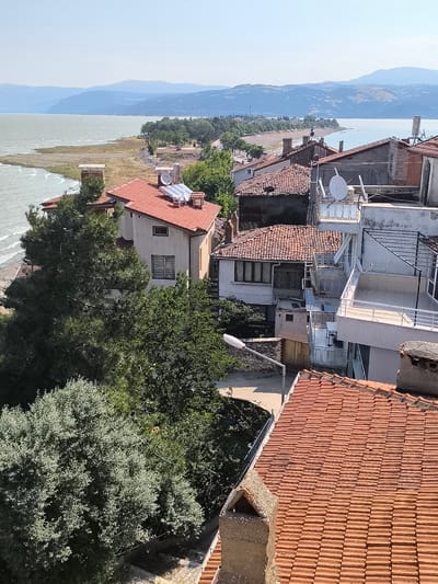 The image captures a scenic view of the city of Eğirdir, Turkey, overlooking the Eğirdir Lake. In the foreground, the terracotta tiled roofs of several houses are visible, with one prominent chimney made of weathered stone. Lush green trees partially obscure some of the buildings. Several houses feature solar panels on their roofs, suggesting a reliance on renewable energy.
Beyond the houses, the expansive Eğirdir Lake stretches towards the horizon, its waters a calm, pale blue under a clear sky. The shoreline is lined with a narrow strip of dry vegetation and scattered trees, indicating a warm, dry climate. In the distance, rolling hills and mountains form a gentle, undulating backdrop against the hazy sky. The overall impression is one of a peaceful, sun-drenched lakeside town.