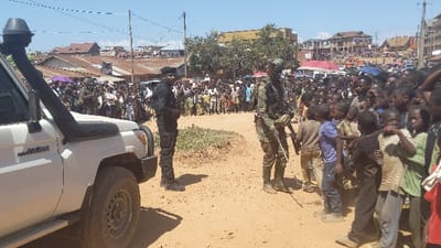 A crowd of people, including many children, are gathered in an outdoor setting. Two uniformed individuals, one in dark attire and another in camouflage, stand as if keeping order. A white SUV is partially visible on the left. The scene seems to be on a dirt road with buildings and additional people in the background. The day is bright, and the atmosphere seems potentially tense with the presence of armed personnel.