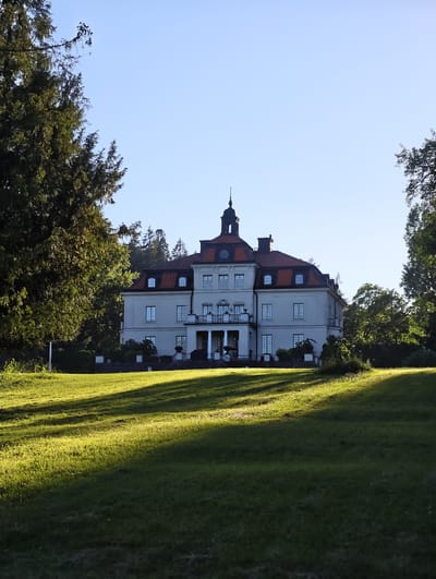 The image shows a large, light-colored building with a red-tiled roof, situated on a grassy slope. The building appears to be a mansion or a large house, with multiple windows and a prominent central tower topped with a cupola. Trees, some dense and dark green, flank the building on either side. The foreground is a sunlit grassy area with strong shadows cast across it, indicating a low sun angle, likely from early morning or late afternoon. The sky is clear and light blue. The location is identified as Skriketorp, Sweden. There are no people visible in the image.