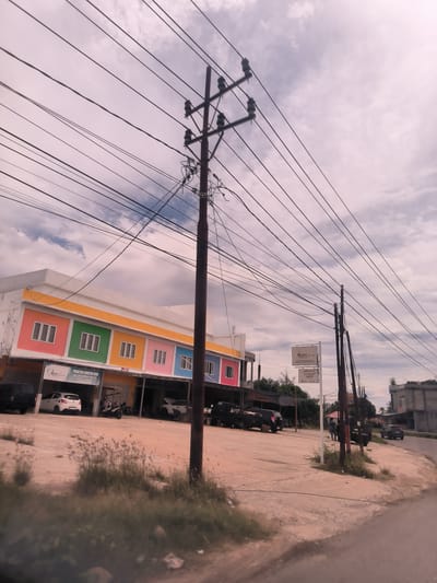The image presents a daytime street scene in Paya Tringgadeng, Indonesia, characterized by a complex network of power lines crisscrossing a cloudy sky. A prominent dark brown utility pole stands centrally in the midground, laden with wires.

To the left, a long commercial building features a vibrant, multi-colored facade with sections of pink, green, yellow, and blue, each with white-framed windows. Signs on this building clearly indicate "Nutri Family Dental Care" and "PRAKTEK DOKTER GIGI" (Dental Practice) with the name "drg. SRI SUHERTI MALATI" and a phone number "0852 1111 8000." Parked in front of this building are several vehicles, including a white hatchback, a black SUV, and a motorcycle.

The immediate foreground shows a patch of dry grass and dirt, transitioning into a paved area where the utility poles are situated. To the right of the main pole, another white sign, partially obscured, also displays "Nutri Family Dental Care." The road curves gently to the right, leading past more utility poles and some distant, more industrial-looking buildings. A black pickup truck, with "MITSUBISHI" visible on its tailgate, is seen further down the road, and a couple of people appear to be standing or walking in that area.

The weather is overcast with light grey clouds. The overall scene suggests a busy local commercial or residential street.