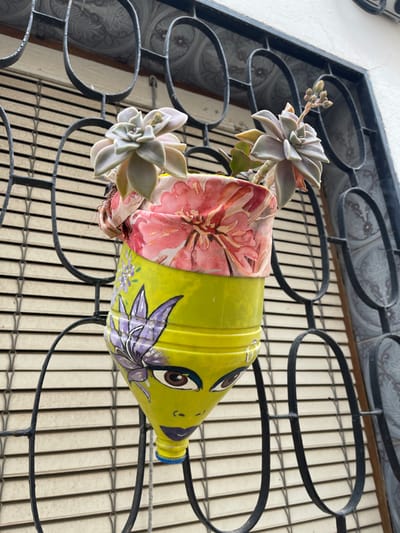 An inverted, repurposed plastic bottle, painted bright lime green, serves as a planter hanging from a black metal grate in Buñol, Spain. The bottle's conical base, pointing downwards, is painted with two wide-set brown eyes, dark pupils, white highlights, and a small purple mouth. Above the eyes, purple and green stylized floral patterns adorn the bottle's side, with small light blue flower dots towards the top left. A patterned fabric strip, featuring large pink, red, and orange floral designs on a light background, is wrapped around the bottle's cut upper rim, partially covering the green paint. Inside, several succulent plants, characterized by thick, rosette-shaped leaves in shades of pale green and grey-purple, are visible. One succulent on the right displays a developing flower stalk with multiple small buds. The black metal grate, composed of vertically aligned oval loops and connecting bars, is positioned against a light-colored window or door covered by horizontal blinds or shutters. A plain white wall is visible in the upper background, with faint patterned tiling or texture to the far right. No individuals are present, and no active interactions are depicted.
