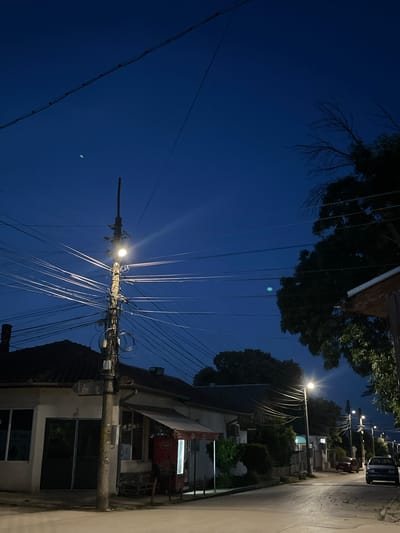 The image depicts a nighttime scene in Dulovo, Bulgaria, characterized by a deep blue twilight sky, trees silhouetted against the fading light, and a street with buildings and utility poles. A single streetlamp illuminates a complex network of overhead wires, casting long, bright streaks of light. The wires are a dominant visual element, crisscrossing the frame and attached to a tall pole.
In the foreground, a building with dark windows and a sloped roof is visible. Further down the street, more buildings line the side, with some appearing to be shops or residences. A car is parked on the street, and another, possibly red, vehicle is partially visible behind it. Trees are scattered throughout the scene, some with dense foliage and others bare-branched. A faint, almost moon-like object is visible in the sky.
The overall atmosphere is quiet and somewhat urban, with the artificial light of the streetlamp and building windows contrasting with the natural twilight. The image captures a typical evening in a residential or mixed-use area. No people are clearly visible, and no discernible text is present in the image. The time of day is clearly night or late twilight, and the weather appears clear.