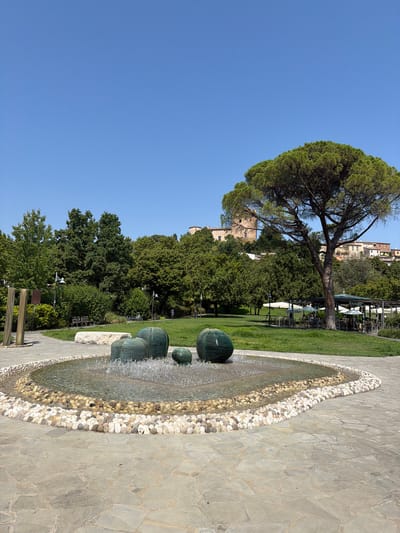 The image depicts a public park in Santarcangelo di Romagna, Italy, on a clear, sunny day. In the foreground, a circular fountain is visible, filled with water and surrounded by a border of various colored pebbles. Several large, green, spherical sculptures are placed within the fountain, with water spraying from them. The fountain is situated on a paved area made of irregular stone slabs. Behind the fountain, a lush green lawn stretches towards a backdrop of trees.
Further in the background, a shaded seating area with tables and umbrellas, presumably an outdoor cafe or restaurant, is set up on the grass. Beyond this, a dense collection of trees forms a natural screen. On a hill overlooking the park, buildings with terracotta roofs and a medieval tower are visible, characteristic of the Italian landscape. A large pine tree with a broad canopy stands prominently in the middle ground, near the seating area and to the right of the tower. A single person is seen walking on the grass in the distance, to the right of the image. The sky is a clear, bright blue.