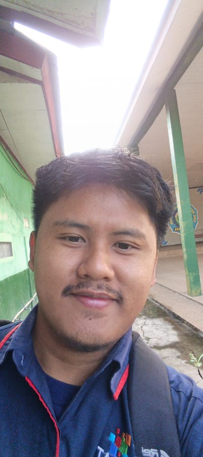 A young, fair-skinned man of Asian descent is captured in a selfie, looking directly at the camera. He has dark, wavy hair and a faint mustache and goatee. He is wearing a dark blue polo shirt with red trim around the collar and a darker shirt underneath. A backpack strap is visible over his right shoulder. The background suggests an outdoor environment with a green wall on the left and a concrete walkway and a green pillar on the right. The lighting is bright and slightly overexposed, indicating it is daytime. The image appears to have been taken in an urban setting, possibly in Sukabumi, Indonesia, given the context of the provided data.