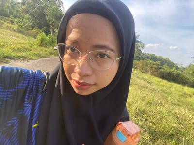 A young woman wearing a black hijab and eyeglasses is taking a selfie outdoors in a grassy, hilly area. She is wearing a black and blue patterned shirt. She holds an orange and blue water bottle in her right hand. The background shows a road, green trees, and a bright blue sky with some clouds. The location is the Gudang Batu area in Indonesia.