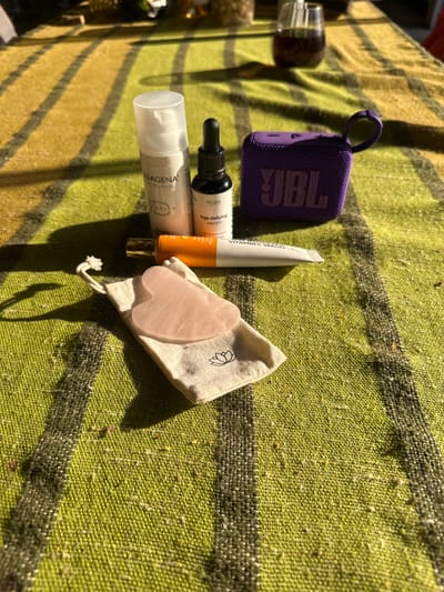 A top-down shot reveals a JBL Bluetooth speaker, skincare products including serums and creams, and a rose quartz Gua Sha tool nestled in a small drawstring bag with a lotus flower design, arranged on a green and tan striped woven blanket. The blanket is laid out in sunlight, likely outdoors, possibly in a city (Pliska, Bulgaria is mentioned, suggesting the location is within that city).