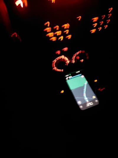 A dark, blurry image shows the interior of a car at night. Illuminated elements include a smartphone displaying a map application, faint red circular rings, and numerous small, blurry red shapes resembling chevrons or stylized "C"s. These lights are likely indicators or part of the dashboard display. The overall impression is one of motion and limited visibility.