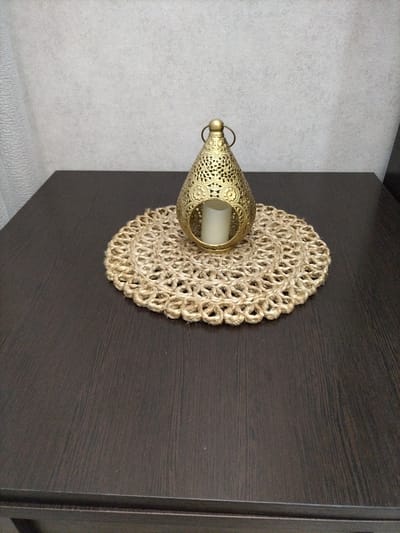 The image shows a close-up, eye-level shot of a decorative brass lantern with a candle inside, placed on a jute placemat. The placemat is circular and has a woven, textured pattern. The lantern itself is ornate, featuring cut-out designs and a small handle at the top. It is positioned slightly off-center on the placemat, which rests on a dark, wooden surface, likely a table. The background consists of a textured, light-colored wall, with a hint of a white curtain on the left side of the frame. The lighting appears to be indoor, with soft illumination that highlights the metallic sheen of the lantern and the natural fibers of the placemat. No people are visible in the image, and there are no discernible signs of text. The overall impression is one of home decor, suggesting a quiet and perhaps cozy atmosphere. The context provided places this scene in Nizhnyaya Makhtama, Russia.