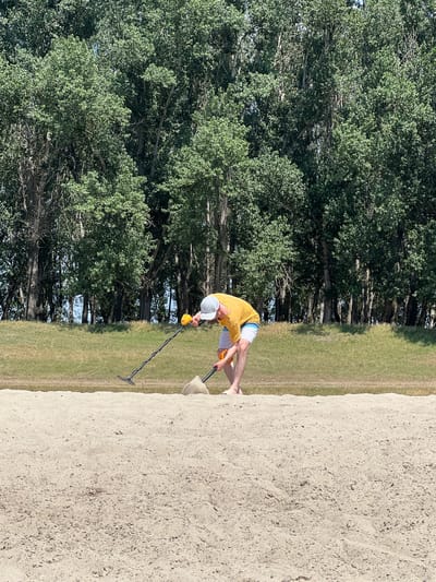 In the city of Indiya, Moldova, a man is metal detecting on a sandy bank with trees in the background. The man, wearing a yellow shirt and white shorts, is bent over and holding a metal detector. A shovel is visible nearby, with a pile of sand next to it, suggesting recent digging activity. The environment is characterized by a clear sky and lush green trees.