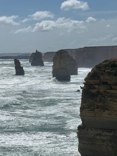 The image captures a coastal scene with prominent rock formations rising from the sea. Rough waves crash against the base of these formations, creating white foamy patterns. In the distance, a coastline or cliff edge is visible under a cloudy sky with patches of blue. The scene is framed by a rocky outcrop in the foreground, adding depth to the composition. The overall atmosphere is one of rugged natural beauty and the power of the ocean.