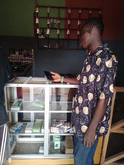 In a mobile phone shop in the city of Balewa, Nigeria, a young man in a black shirt with a patterned design stands by a glass display case. The shop has green and red walls. The display case is filled with various mobile phones and accessories. On a shelf above the case, there are more mobile phones displayed on a wooden board. The man is holding a dark-colored smartphone in his right hand. He is looking down at the phone. The image appears to be taken indoors, possibly during the daytime.