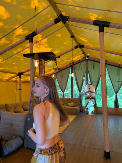 The image depicts the interior of a large, yellow tent structure with wooden poles and a fabric ceiling. A young woman with long brown hair, featuring streaks of pink and blonde, is shown in profile facing left, wearing a light yellow crop top and a patterned skirt. She has a silver earring in her left ear. In the background, there are beige sofas, a tall outdoor heater, and several hanging light bulbs. The tent's back wall appears to be made of draped fabric, with an opening revealing a view of green trees. A full-length mirror is positioned on the floor in the middle of the space, reflecting the interior and the outside environment. The floor is made of wooden planks. The scene appears to be an outdoor glamping accommodation located in a natural setting, possibly within or near the city of Pod'yapol'skoye, Russia.