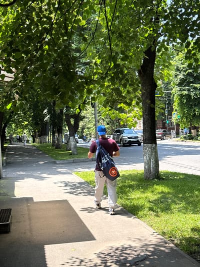In the city of Ver. Alkun, Russia, a person wearing a maroon t-shirt, light-colored pants, a blue baseball cap, and carrying a dark blue backpack with an orange circular emblem, walks on a paved sidewalk. The sidewalk is lined with green grass and trees that cast dappled shadows on the ground. In the background, cars are visible on a street, and buildings are present on the other side of the street. The overall scene depicts a sunny day in an urban environment with pedestrian activity.