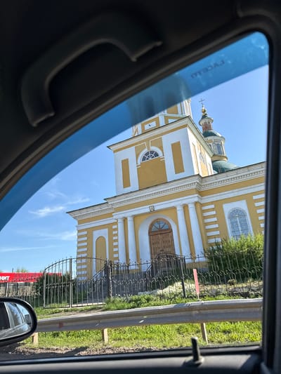View of the Church of the Intercession of the Holy Virgin in the village of Chutyr, Russia, as seen through a car window. The church is yellow and white, with a dome and cross visible against a blue sky. A fence separates the church from a grassy area. Part of a guardrail is visible. A red sign with Cyrillic text is visible in the side mirror.