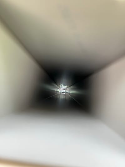 An out-of-focus image shows a dark, square-like tunnel illuminated by light at the far end. The light reflects off what appears to be some sort of metal grid. The tunnel's walls are blurry and white, creating a sense of depth and distance. The framing suggests a perspective looking directly into the tunnel.