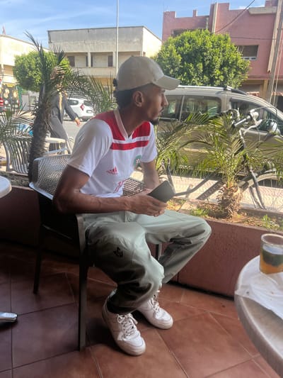 A male individual is seated outdoors at a cafe in Mechra Bel Ksiri, Morocco, on a sunny day. He wears a beige baseball cap, a white t-shirt with red stripes and an emblem (likely the Moroccan crest), light green cargo pants with a dark logo on the left leg, and white sneakers. His hair is dark and styled in a small bun under his cap, and he has light facial hair. He holds a dark-colored smartphone with both hands, looking towards the right side of the frame. To his right, on a light-colored round table, a glass of yellowish-orange liquid with a mint sprig (presumably mint tea) and a crumpled white napkin are present. The ground is covered in reddish-brown square tiles. In the background, a street scene unfolds with multiple palm trees separating the cafe area from the road. A person in dark attire walks across the street from left to right. A silver SUV is parked further down the street, and parts of a black bicycle with the word "Peugeot" on its frame are visible. Across the street, buildings with weathered light grey and reddish-pink facades are visible under a clear blue sky.