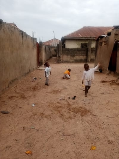 The image depicts three children in an outdoor alley in a Nigerian location. The alley is defined by high, weathered walls on either side, leading towards houses with corrugated iron roofs. The ground is a sandy, reddish-brown color, with scattered debris. One child, wearing a white shirt and shorts, stands in the foreground with an arm raised. Another child is crawling or kneeling on the ground, wearing an orange shirt. The third child stands further back, wearing a white t-shirt and looking towards the camera. The sky is overcast. The location is characterized by basic construction and general untidiness.