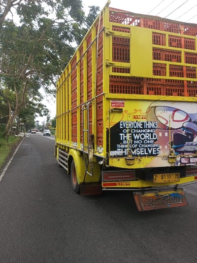 This image depicts a brightly colored yellow truck, configured as a livestock carrier, positioned on the left side of an asphalt road in the city of Kamji Na-Yekha, Bhutan. The truck's cargo bed features a multi-tiered, cage-like structure entirely filled with numerous orange plastic crates. The rear of the truck displays a yellow bumper, red rectangular taillights, and a license plate marked 'Z 9110 FD' with '09-26' visible below. A prominent black sticker on the tailgate bears the quote, "EVERYONE THINKS OF CHANGING THE WORLD, BUT NO ONE THINKS OF CHANGING THEMSELVES," alongside a stylized graphic depicting an anime-like character with a masked helmet and large eyes. A smaller yellow sticker on the truck's side includes a phone number, '0823-3777-3776,' and partially visible text. Below the license plate, a decorative wooden or metal mudflap is painted with the word 'Realistica' in a stylized script. A metal ladder is attached to the rear-left side of the truck bed. The road is bordered by a grassy shoulder and mature green trees under an overcast sky. In the background, several vehicles, including white cars, are visible traveling on the road. No individuals are visible within the frame.