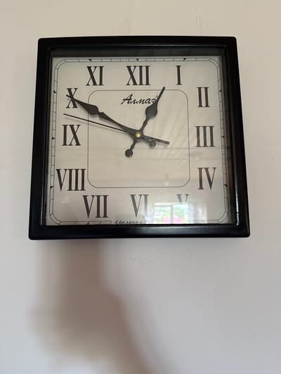A square, black-framed clock with a white face and Roman numerals is mounted on a white wall. The clock has black hour and minute hands. The brand name "Armaz" is visible on the clock face. Text that appears to be Russian is located at the bottom of the clock face. A shadow is cast on the wall below the clock. The image's metadata indicates it was taken in Khapo-Oye, Russia, which is a village in the Republic of Buryatia.