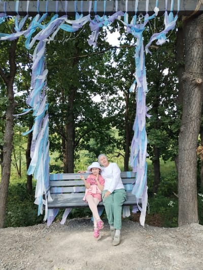 Two people, likely an adult and a child, are sitting on a wooden bench suspended from a swing-like structure. The structure is adorned with streamers in shades of white, blue, and purple, creating a festive atmosphere. The child, wearing a pink t-shirt, shorts, and a white hat, is seated on the left side of the bench. The adult, dressed in a white long-sleeved shirt and green pants, is sitting next to the child on the right. Both appear relaxed and comfortable.
The scene is set outdoors, with a backdrop of lush green trees, suggesting a park or natural setting. The ground around the bench is covered in gravel. The lighting suggests it is daytime, possibly late morning or afternoon, with ample natural light. The weather appears pleasant, with no indication of rain or extreme conditions. The presence of the decorated swing implies a recreational or celebratory event might be taking place, or it's a dedicated photo spot.
The t-shirt of the child has visible text that reads "REAL". The overall mood conveyed by the image is one of peacefulness and enjoyment. The location context provided, Blokhino, Russia, places the scene within a specific geographical area.