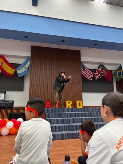 Scene in Tlaxcalancingo, Mexico: A man in a hat and jacket stands on a stage against a striped wooden backdrop. Above him are flags of Brazil, Portugal, the United Kingdom, and the United States. Below him, flags of Argentina and Spain flank a black monitor. Two children wearing white shirts and red headbands stand on a blue-striped floor. One person in a white shirt stands to the side. Orange balloons are visible in the foreground. The word "ROAR" is displayed in yellow lettering behind the man.