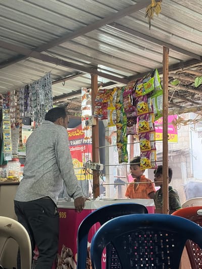 A man in a striped shirt stands in a small shop, tending to a display of snacks and candies hanging from the ceiling. Shelves below are stocked with various goods. Signage in both English and another language (possibly Kannada) is visible, with the phrases "FAMILY HOME" and "Noodle" appearing prominently. A blue plastic chair is in the foreground. The shop seems to be located on a street.