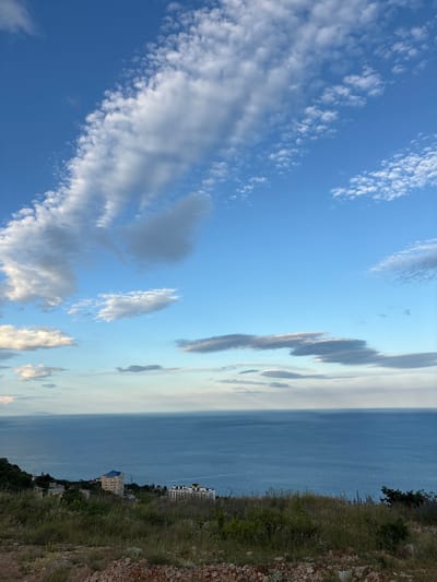 This image depicts a coastal landscape near Malyi Mayak, Ukraine. The scene is dominated by a vast expanse of the Black Sea under a bright blue sky filled with scattered clouds. In the foreground, a grassy hillside slopes down towards the sea, with some exposed rocks and dry vegetation. Along the coastline, several buildings are visible, including apartment complexes and smaller houses, nestled amongst greenery. The water exhibits a deep blue hue, with subtle ripples indicating a calm sea. The sky is a vibrant blue, with wispy white clouds forming various shapes across the upper portion of the image, some appearing thicker and more defined, while others are thinner and more spread out. The overall impression is a serene and picturesque coastal view.