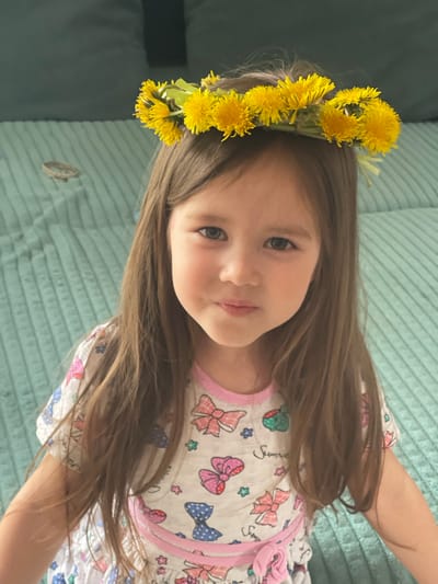 A young girl with long brown hair wears a wreath of yellow dandelions on her head. She is wearing a light-colored top with a pattern of bows and other designs. The background is a textured, turquoise-colored surface. The image was taken in Verkh-Katunskoye, Russia.
