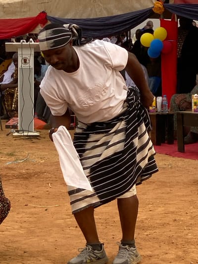 A man stands outdoors on dirt, wearing a white long-sleeved shirt with subtle branding and a black and white striped cloth wrapped around his waist like a skirt. He also sports athletic shoes and a matching striped cap. A white cloth is in his right hand. In the background, a structure with red and blue draped fabric is visible, along with balloons and tables with various items.