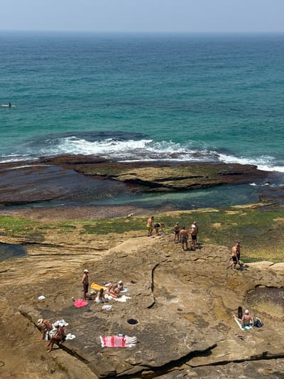 An aerial view captures a rocky coastline with people enjoying the sun. The scene features the ocean with waves crashing against the shore. Individuals are seen swimming, walking along the rocks, and sunbathing on towels. Some are gathered in groups while others are solitary. The landscape is a mix of stone, sand, and vegetation, creating a textured and sun-drenched coastal setting.