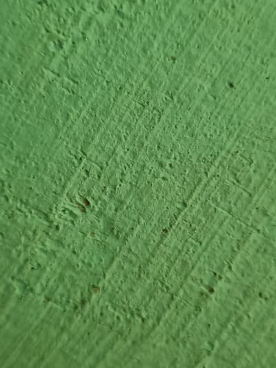 The image is a close-up, abstract view of a green, textured surface. The surface appears to be painted plaster or concrete, with visible trowel marks and small indentations or pores. The lighting suggests an outdoor setting, possibly during the daytime, as indicated by the even illumination across the surface and the absence of harsh shadows. There are no discernible subjects like people or objects, nor any clear scene or setting. The activity is static, focusing solely on the texture and color of the wall. Notable details include the uneven, slightly rough texture and the consistent, vibrant green color. There are no apparent location cues beyond the fact that the image was taken in Žygaičiai, Lithuania. No text is visible in the image. The overall impression is one of a simple, everyday surface detail.